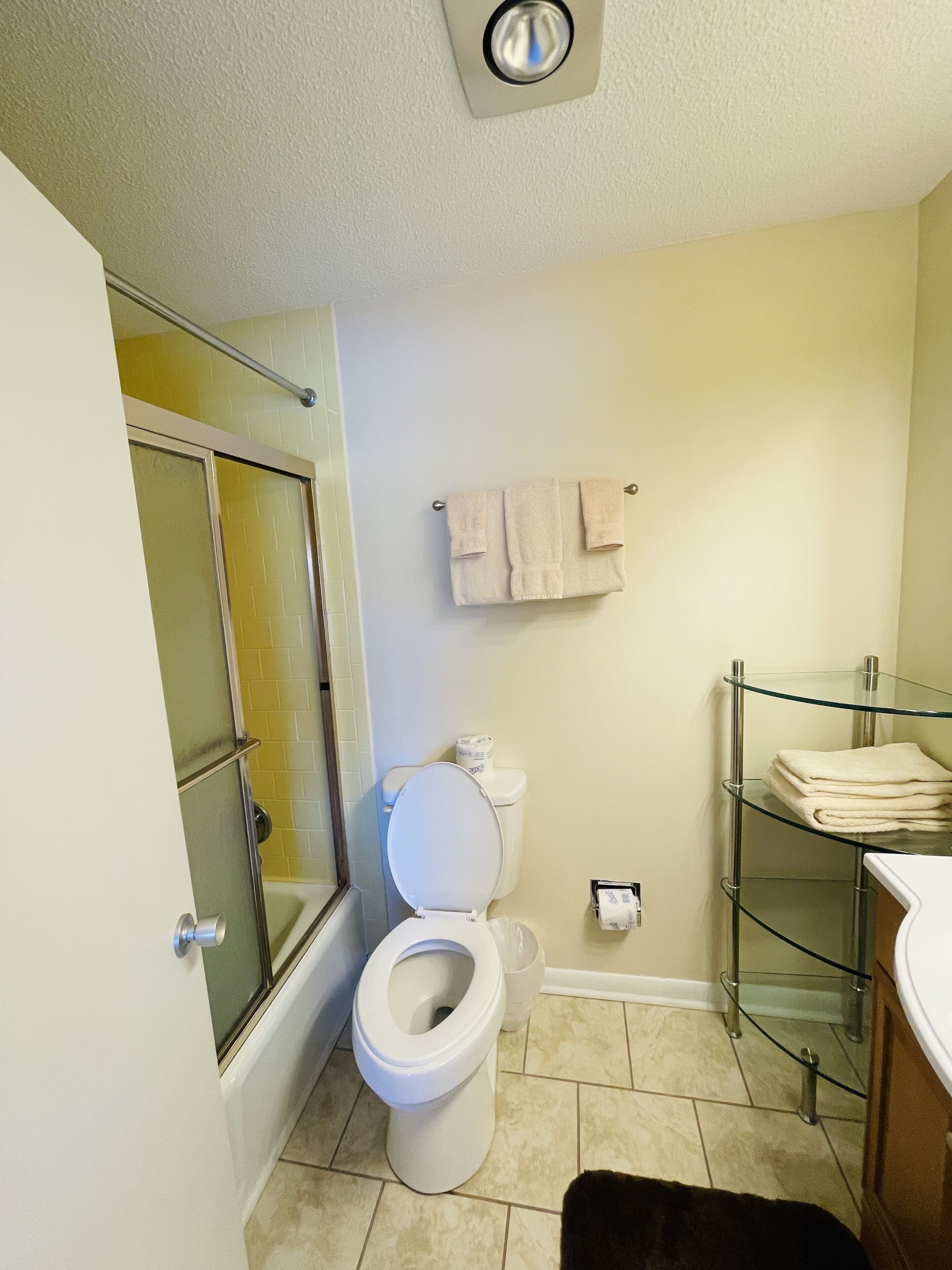 A bathroom with a glass shower enclosure, a toilet, a glass shelf with folded towels, and a white sink.