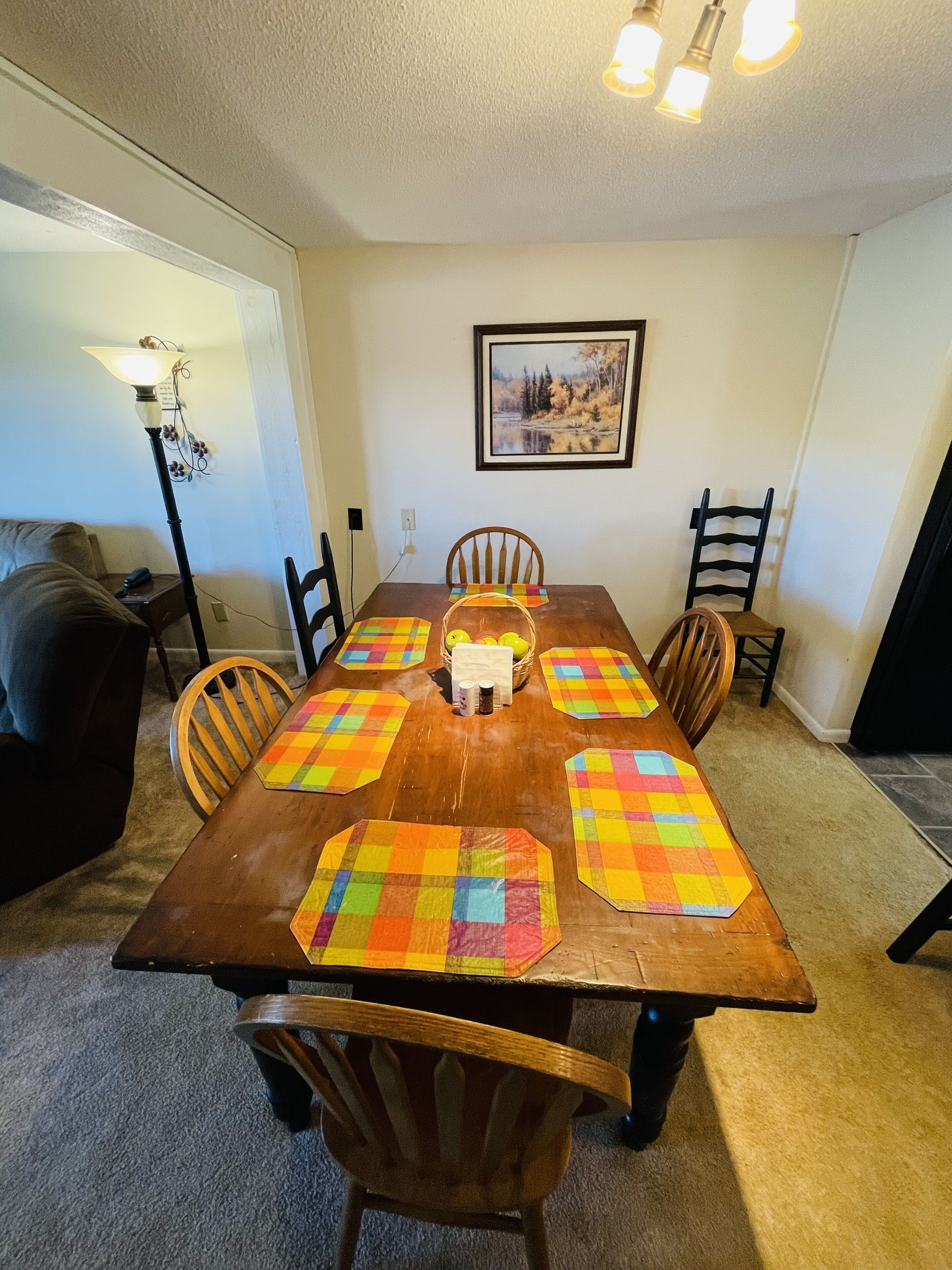 Une salle à manger avec une table en bois, six chaises, un lustre, un tableau encadré au mur et une lampe sur une table d’appoint.