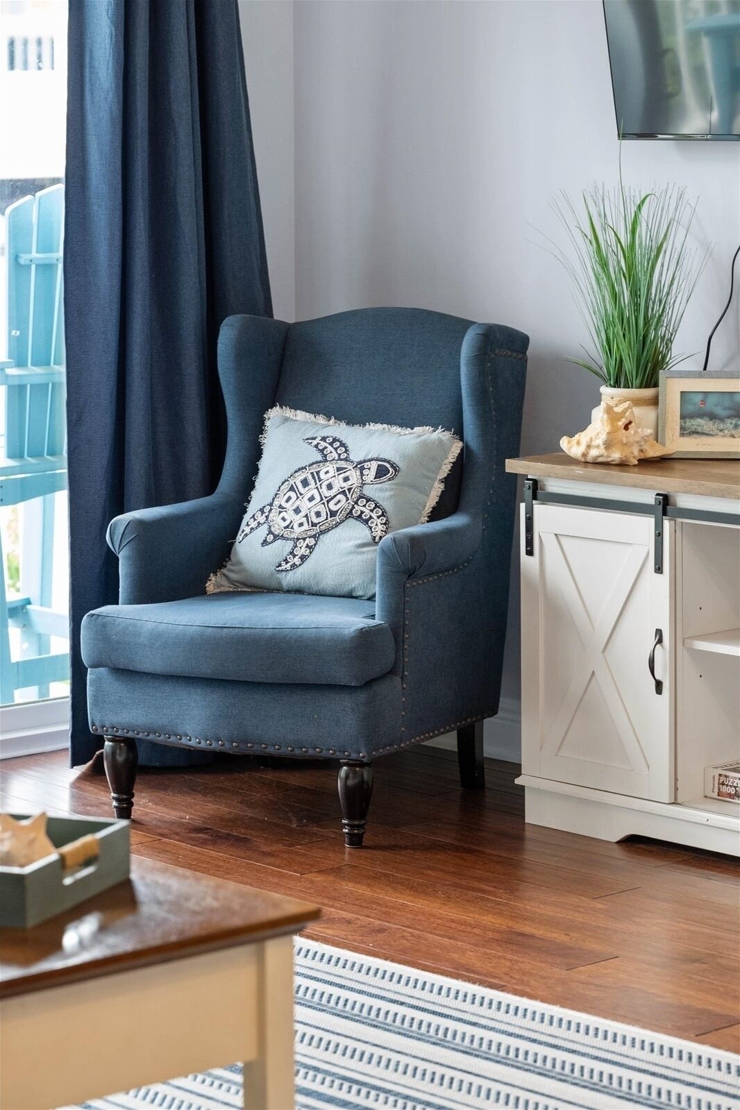 Un fauteuil bleu avec un coussin à motifs, une armoire blanche, une plante et un tableau encadré posés sur une table en bois.