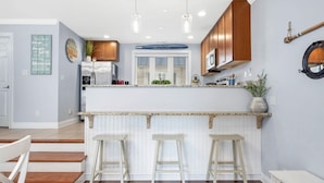 A modern kitchen with wooden cabinets, a central island with stools, and a view of the living area. - Beach Paradise III - DOWNTOWN OCEANVIEW Luxury Townhome-Steps to Beach&Boardwalk (Ocean City)