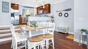 A modern kitchen with white cabinetry, a central island, and a dining area with white chairs and a wooden table. - Beach Paradise III - DOWNTOWN OCEANVIEW Luxury Townhome-Steps to Beach&Boardwalk (Ocean City)
