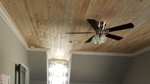 Ein Schlafzimmer mit Holzboden, Deckenventilator, Bett mit Kissen, Nachttisch und Sessel.