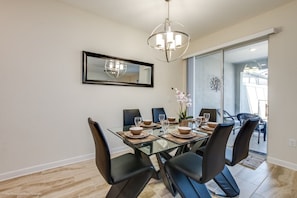 Un coin repas avec une table en verre, des chaises noires et un lustre. Un miroir orne le mur, et une porte coulissante offre une vue sur l’extérieur.