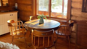 L’intérieur chaleureux d’un chalet comprend une table à manger en bois entourée de chaises, une grande fenêtre donnant sur les arbres et un coin cuisine avec un évier et des placards.