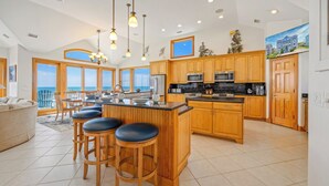A spacious kitchen with wooden cabinets, a central island with bar stools, and large windows offering a view of the ocean. - Luxury Oceanfront Home w/ Pool, HotTub, EV Charger, Elevator, Theater, Much More (Hatteras)