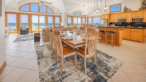 A spacious dining area with a large wooden table and chairs, a kitchen with wooden cabinets, and a view of the ocean through large windows. - Luxury Oceanfront Home w/ Pool, HotTub, EV Charger, Elevator, Theater, Much More (Hatteras)