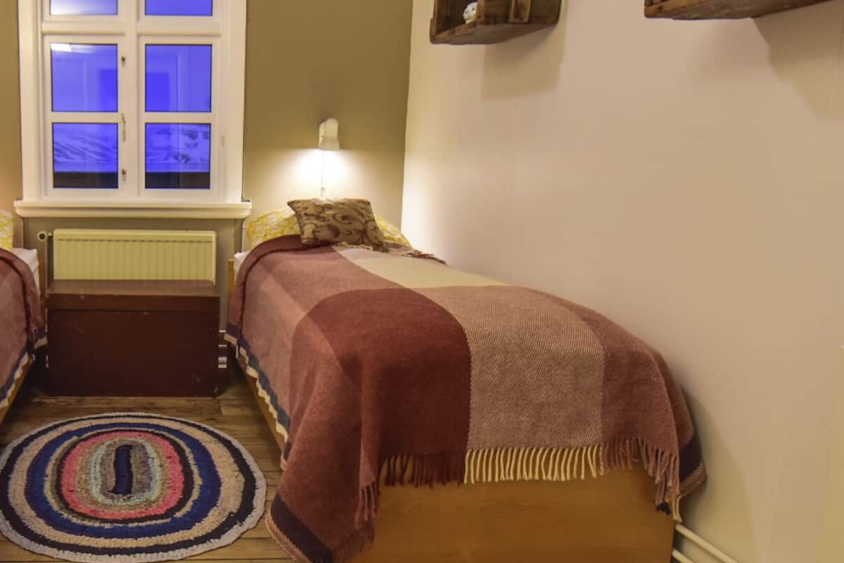 A bedroom with a window, a bedside table, a lamp, a rug, and a shelf with a plant.