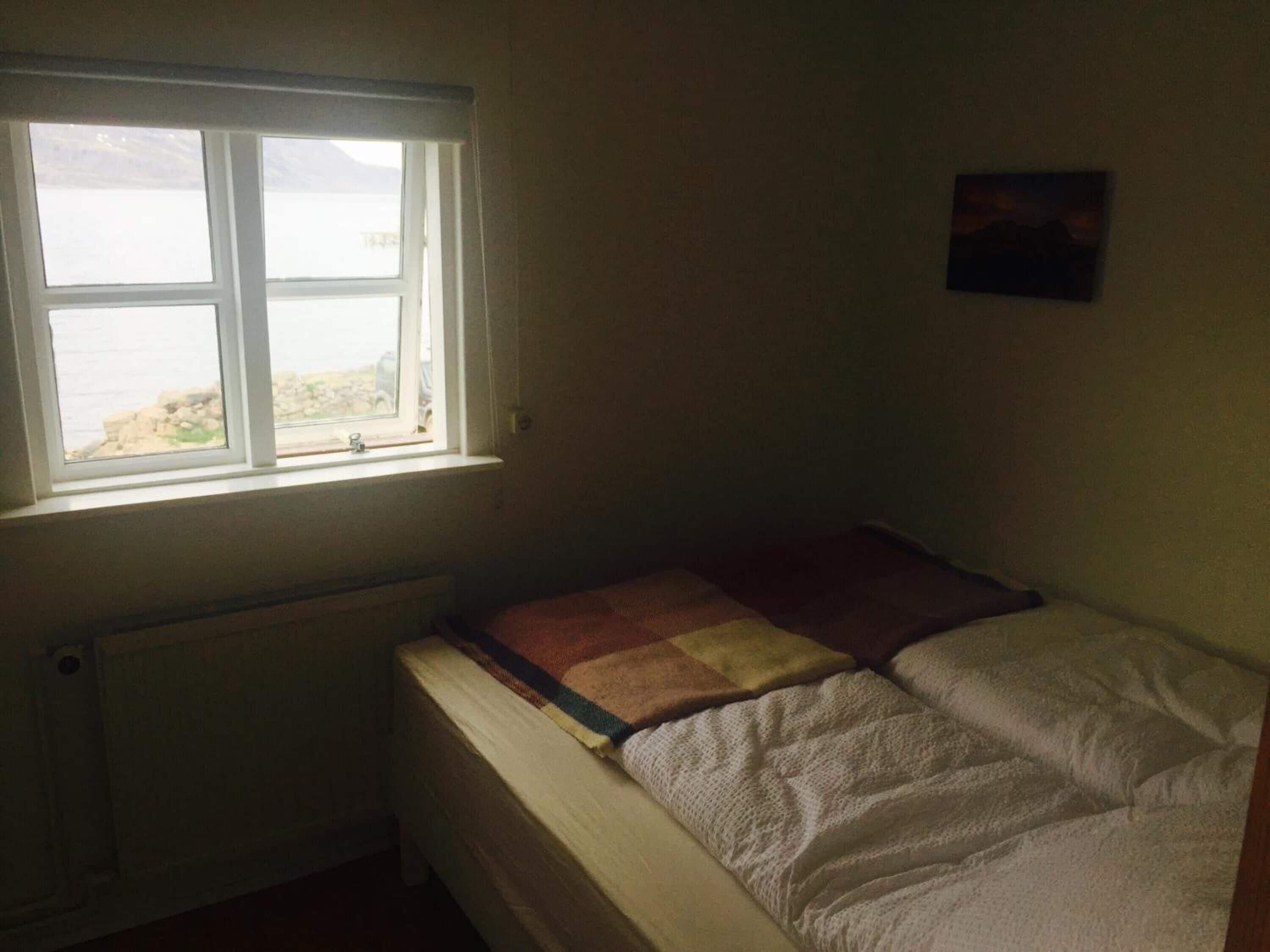 A small, dimly lit room with a single bed, a radiator below the window, and a small painting on the wall.