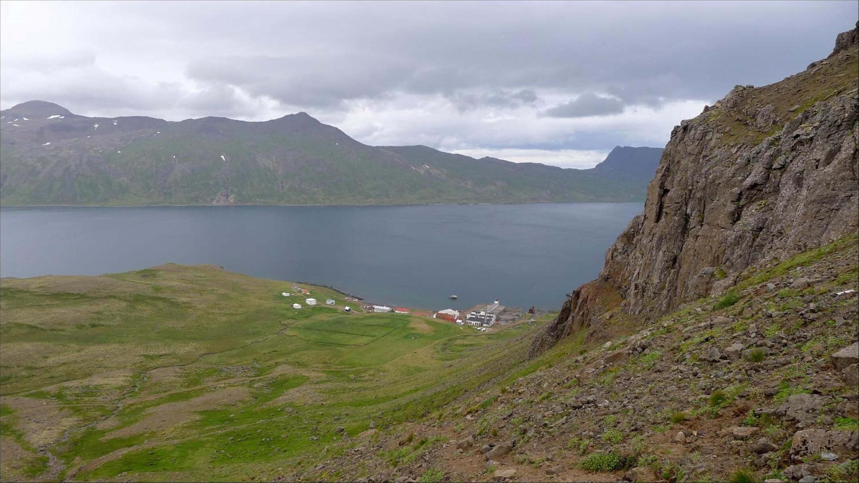 Un paisaje montañoso con un lago, un pequeño poblado con edificios y un acantilado rocoso.