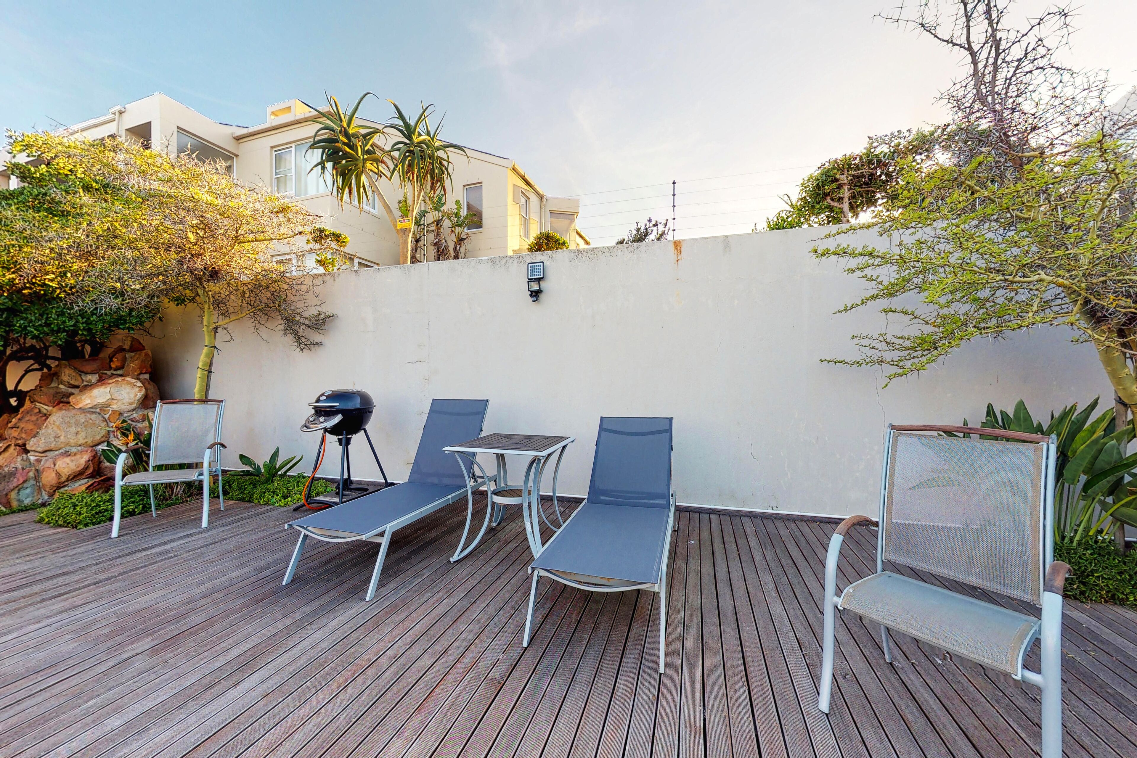 A wooden deck with four lounge chairs and a small table, surrounded by a white wall and greenery.