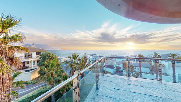 A balcony with a glass railing overlooking a coastal area with palm trees and buildings. - Luxury Beach Villa (Cape Town)