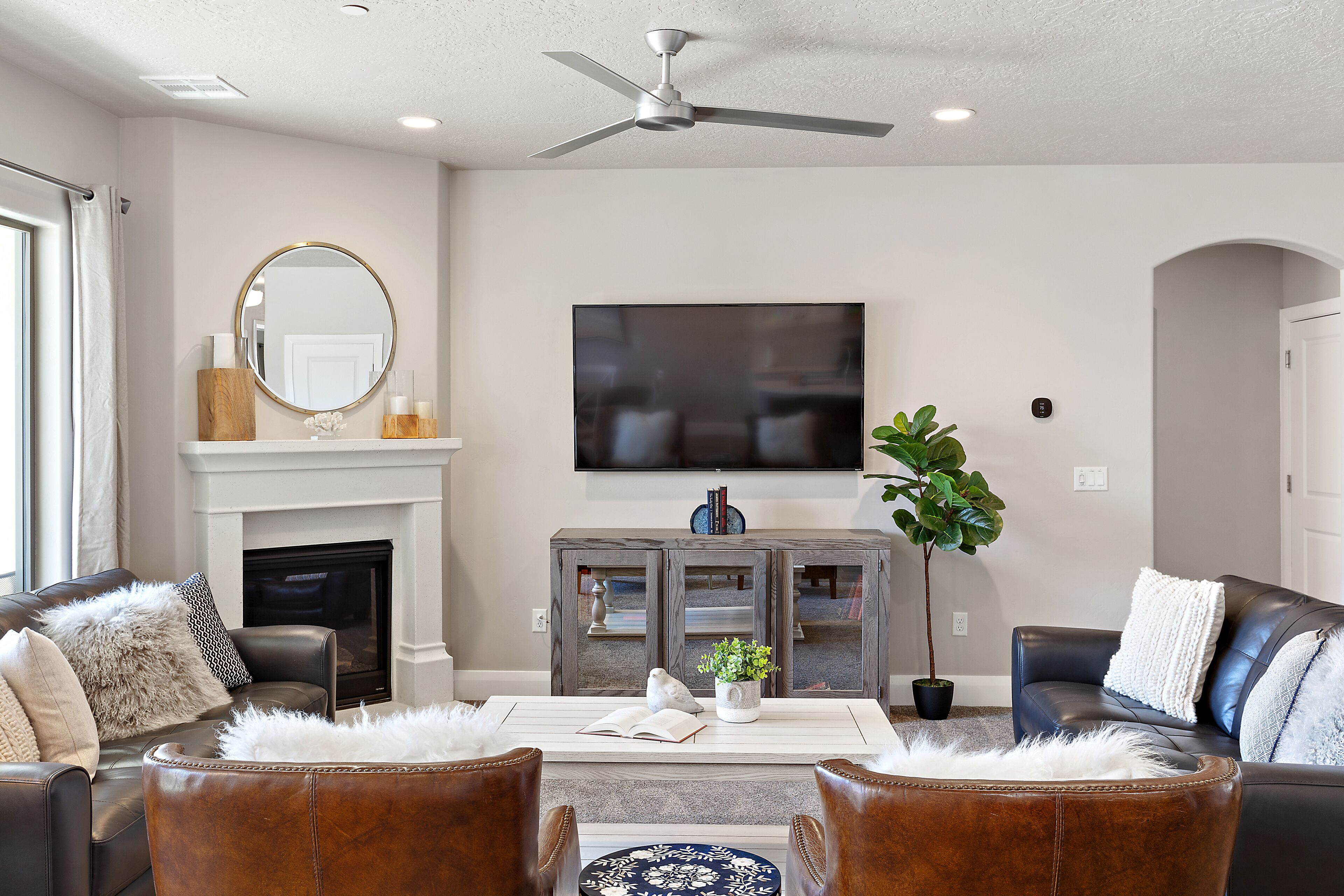 A modern living room with leather sofas, a fireplace, a flat-screen TV, and a ceiling fan.