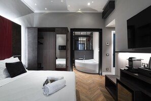 Ein modernes Hotelzimmer mit einem großen Bett, integriertem Fernseher, einem Flachbildfernseher auf einem Tischchen sowie einem Badezimmer mit Badewanne und Holzboden.