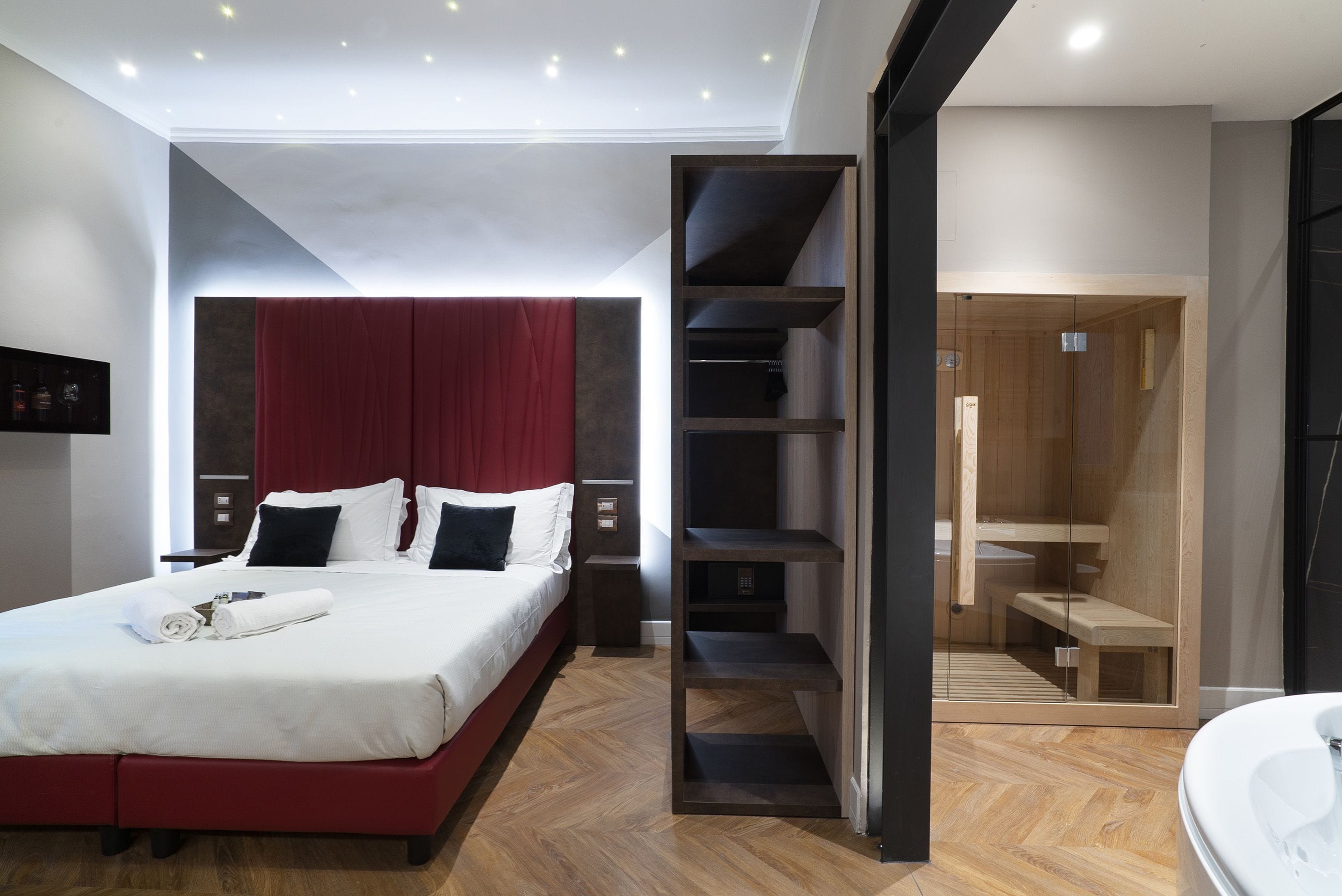 A modern hotel room with a large bed, a red headboard, a built-in wardrobe, a wooden floor, and a separate bathroom area.