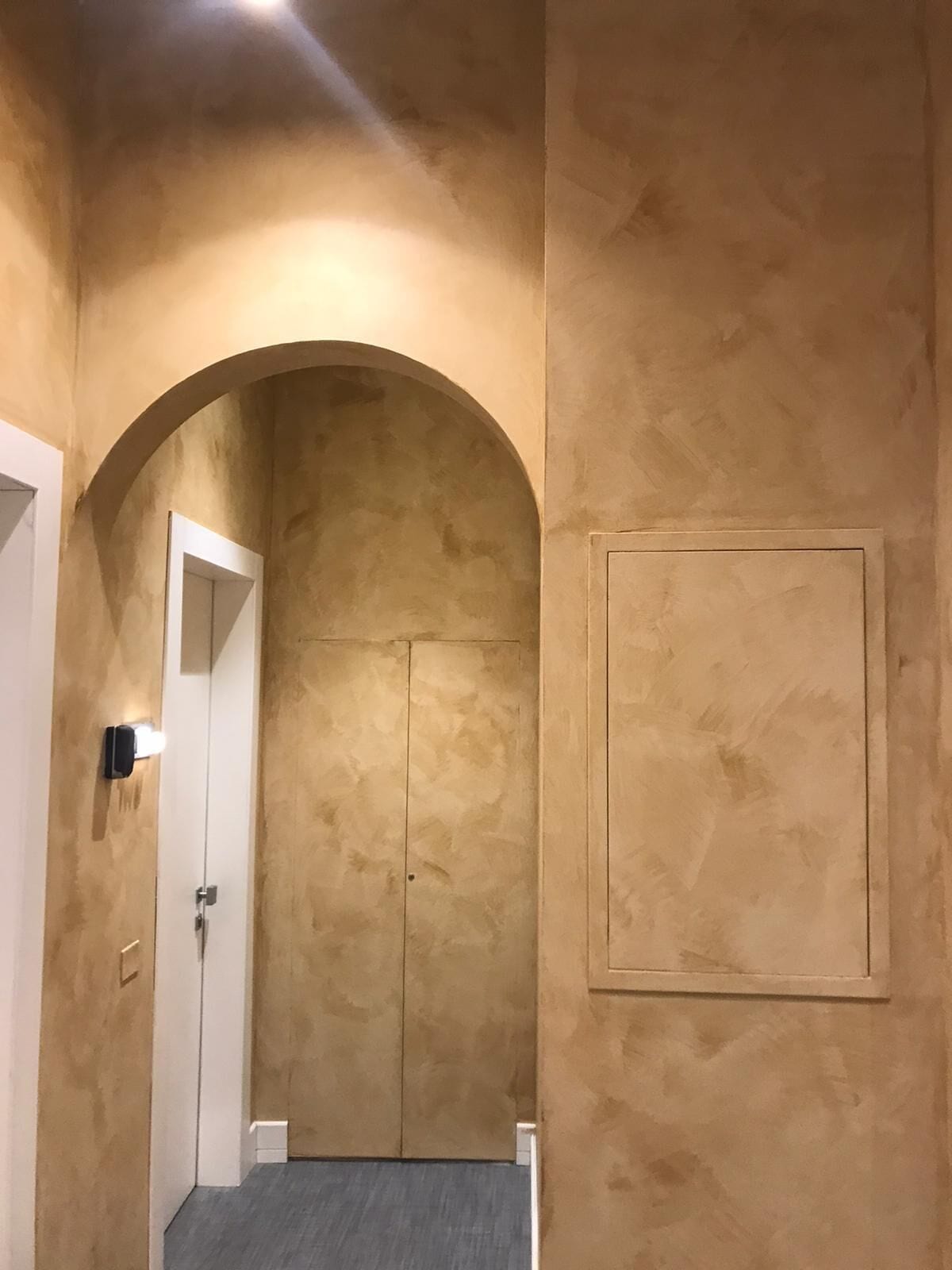 A hallway with a textured beige wall, an arched doorway, and a door with a light switch on the wall.