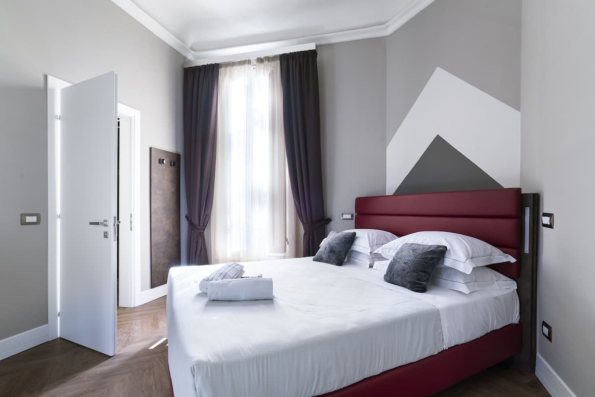 A modern hotel room with a large bed, a red headboard, and a view of the city through the window.