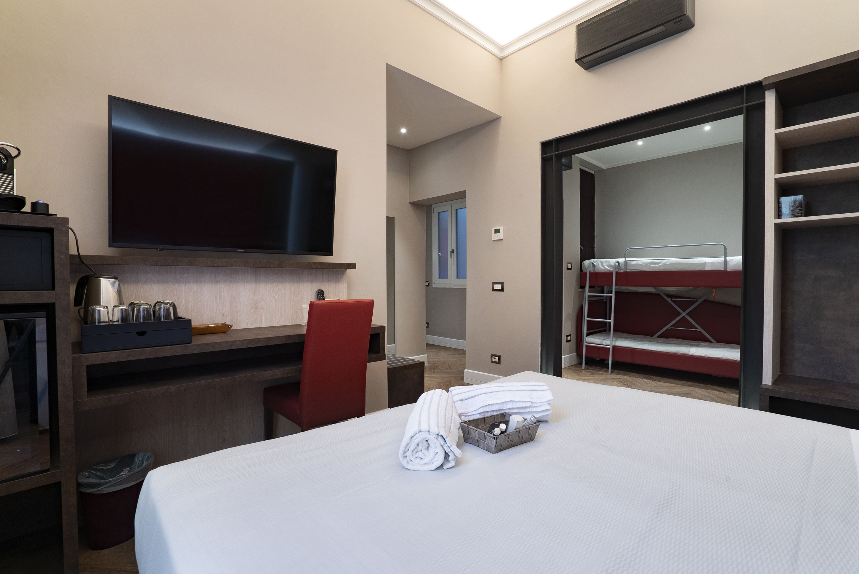 Una camera d'albergo con un letto, una scrivania, una TV, una sedia rossa, un letto a castello e un piccolo balcone.