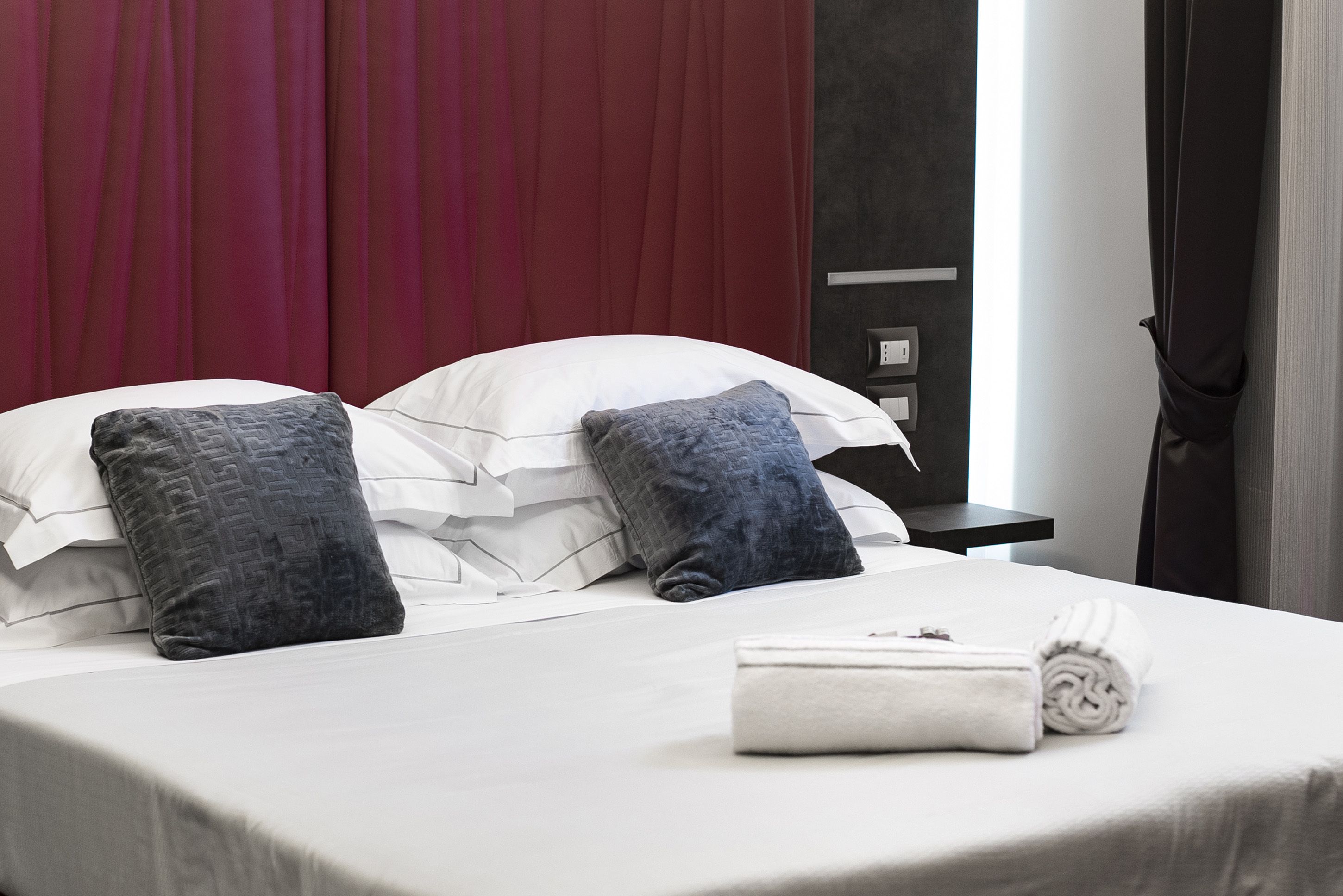 Un lit bien fait, avec du linge de lit blanc et deux oreillers foncés, une serviette pliée et une table de chevet sombre dotée d’une prise électrique.