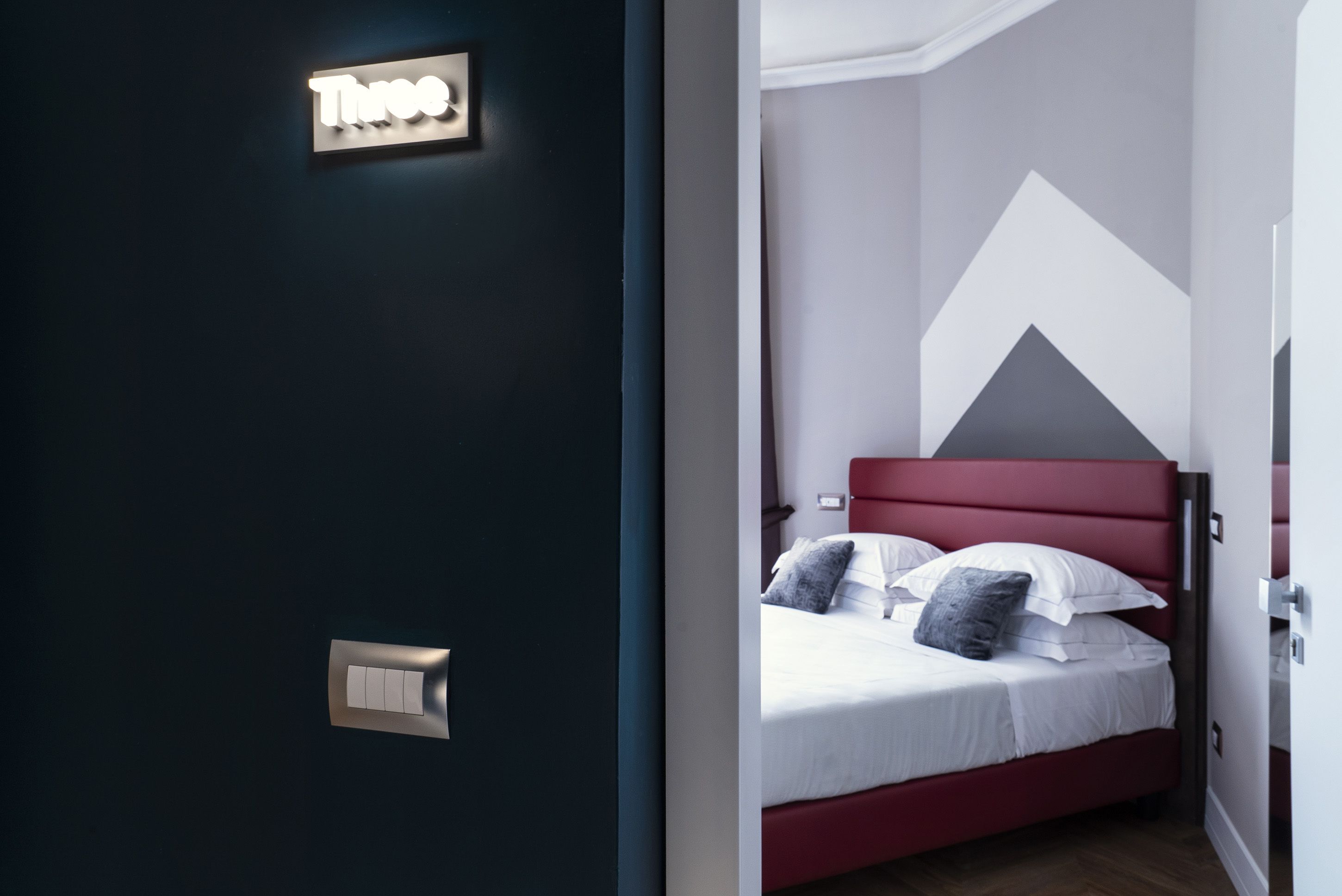 Ein modernes Schlafzimmer mit einem roten Kopfteil, einem Bett mit weißer Bettwäsche und grauen Kissen sowie einem an der Wand montierten Lichtschalter.