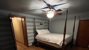 Un dormitorio con cama con dosel, suelo de madera, ventilador de techo y una puerta que da a otra habitación.