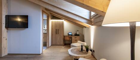 A cozy attic room with a wooden ceiling, a flat-screen TV, a round wooden table with a lamp, and a cozy seating area.