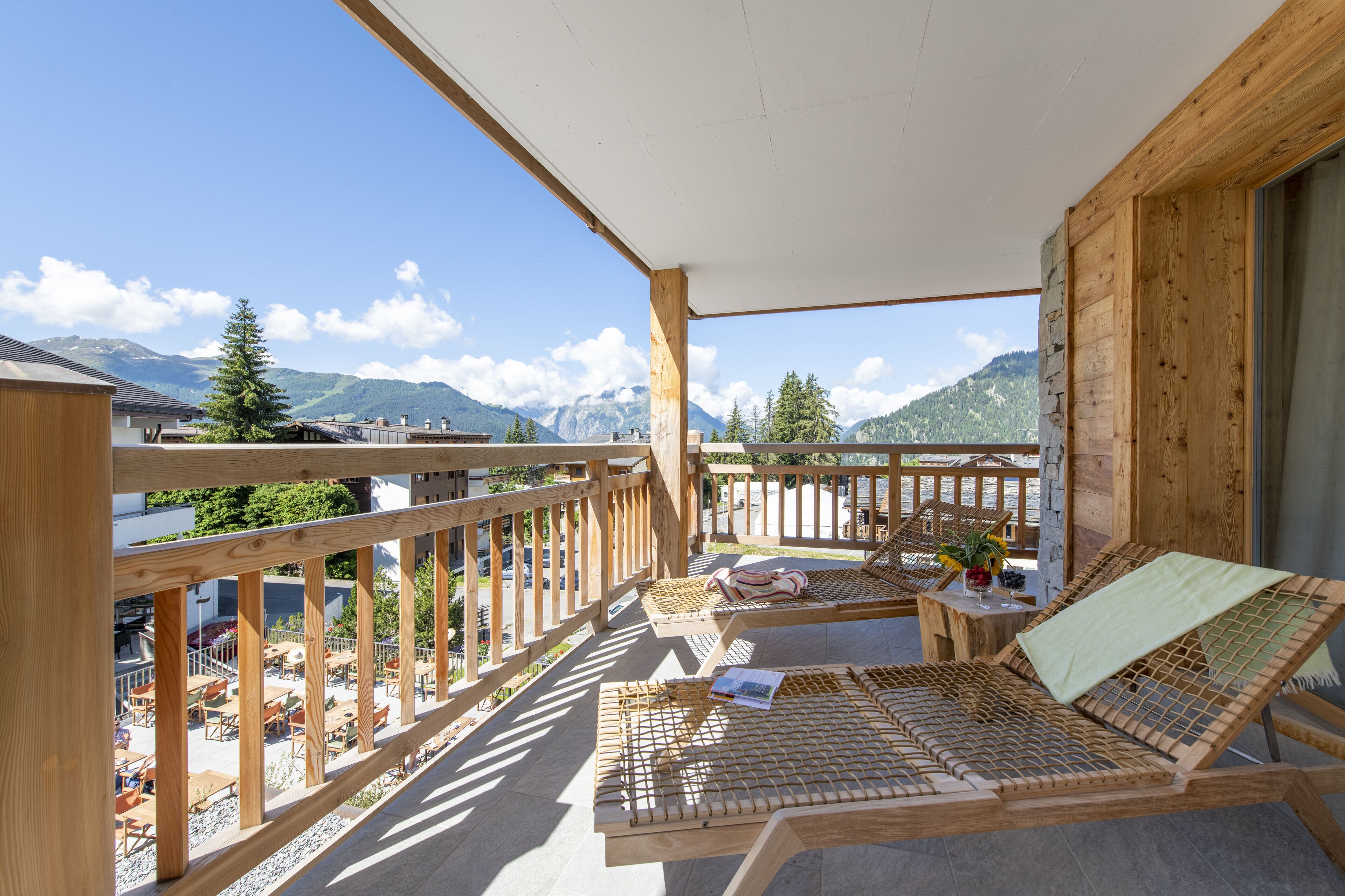 A wooden balcony with a view of mountains, a table with a book, and a lounge chair.
