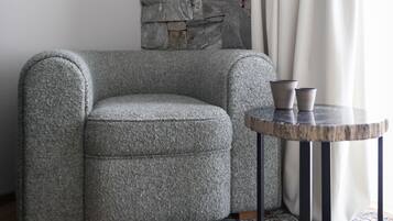 Habitación de esquina con pared de piedra, un sillón gris, una mesa redonda con dos tazas y una alfombra estampada.