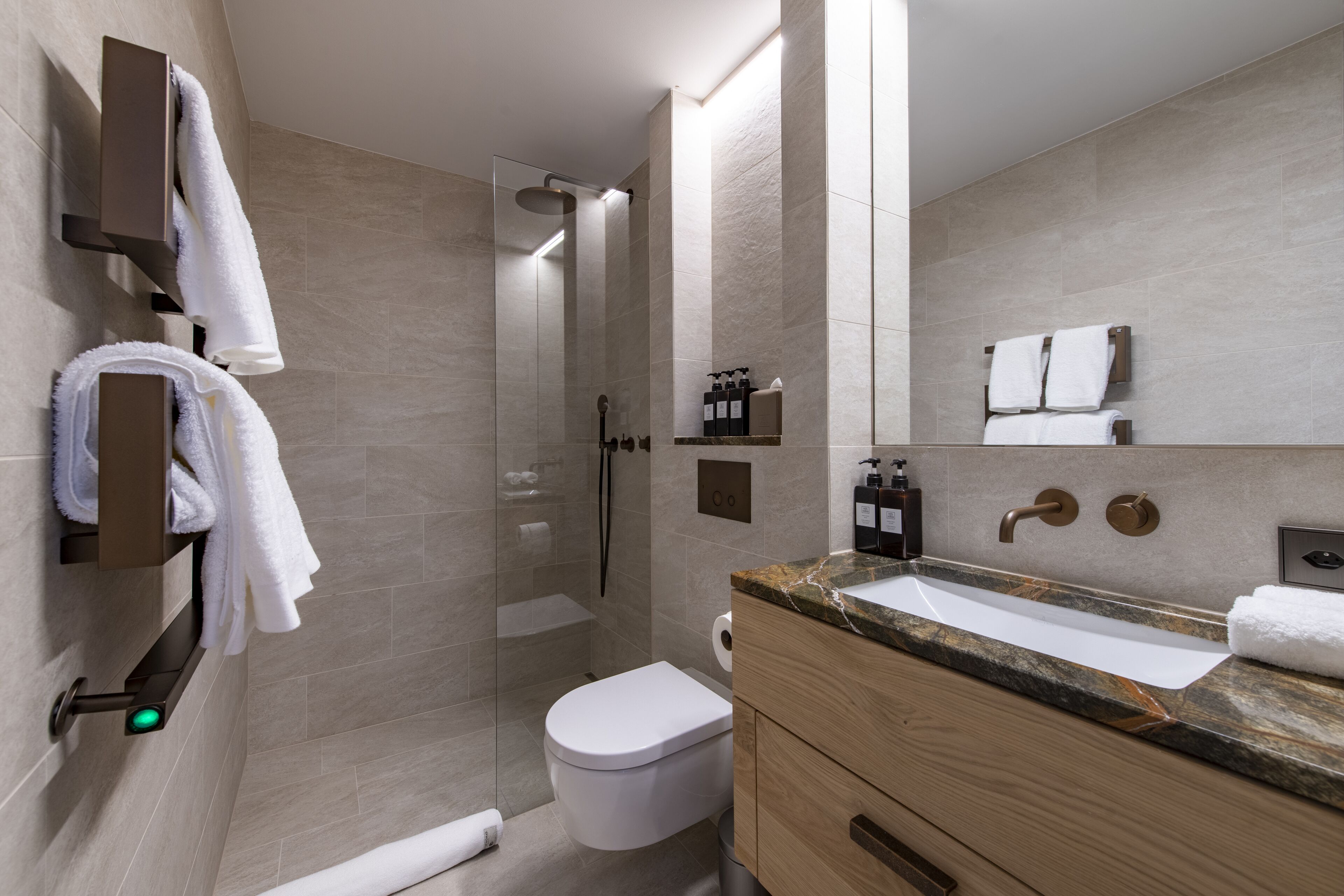 Un baño moderno con ducha envuelta en vidrio, inodoro blanco, lavamanos sobre encimera de mármol y muebles de madera.