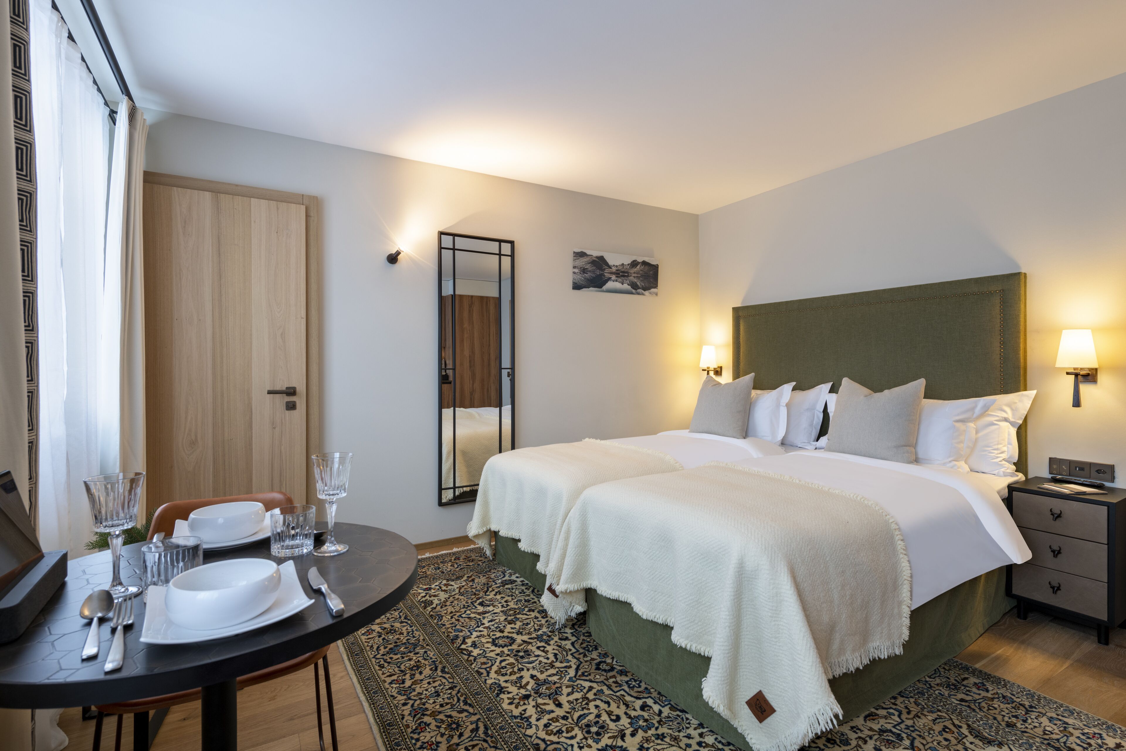 Une chambre d’hôtel avec un lit double, des tables de chevet, un petit bureau avec une chaise, une table à manger avec des couverts et un tapis à motifs.