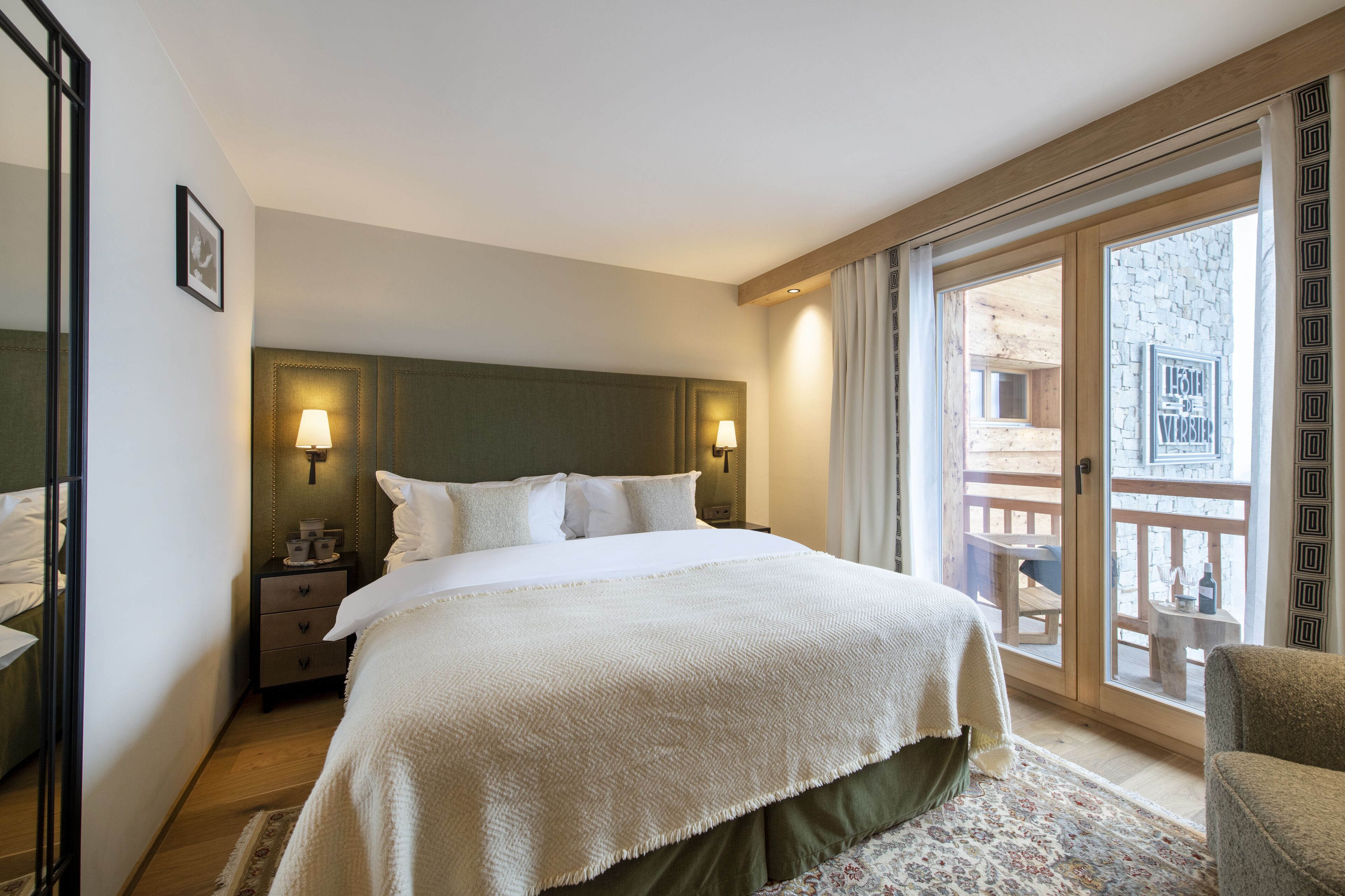 A hotel room with a large bed, two bedside lamps, a nightstand, a chair, and a balcony with a view of the city.