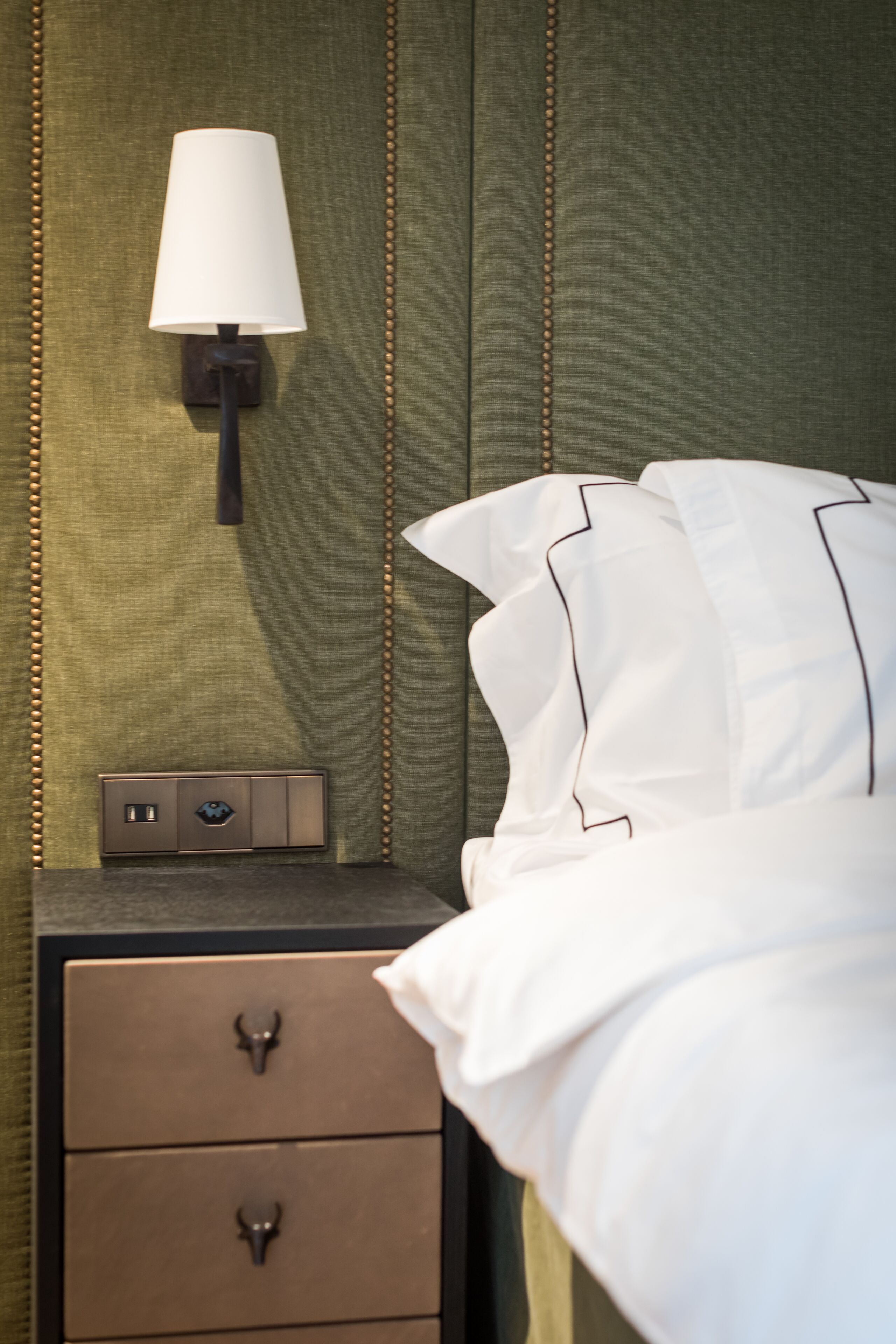 A neatly made bed with white linens and a pillow, a bedside lamp, and a small nightstand with a switch and a decorative item.