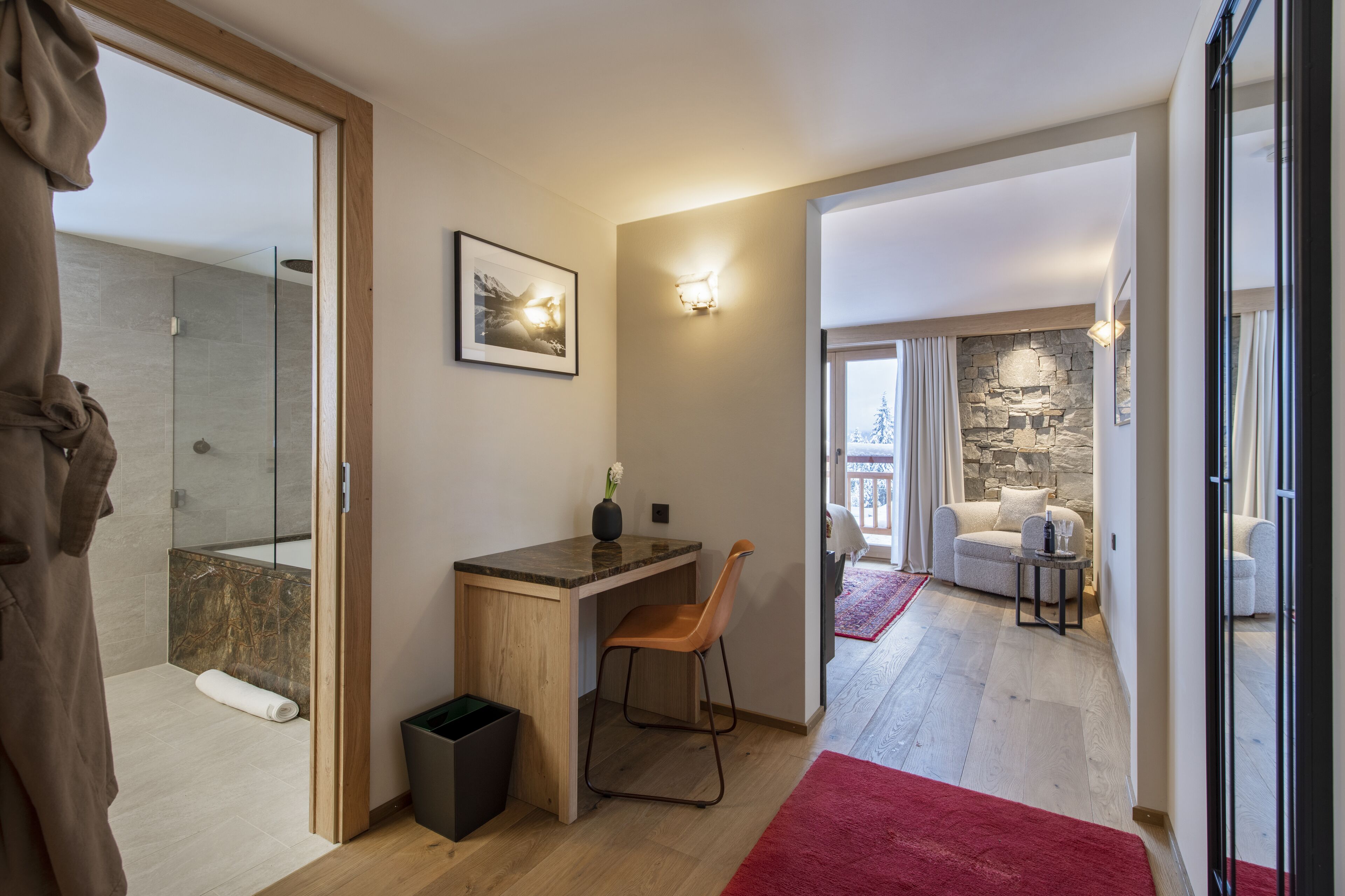 A modern bathroom with a shower, a wooden desk, and a chair. There is a red rug on the floor and a framed picture on the wall.