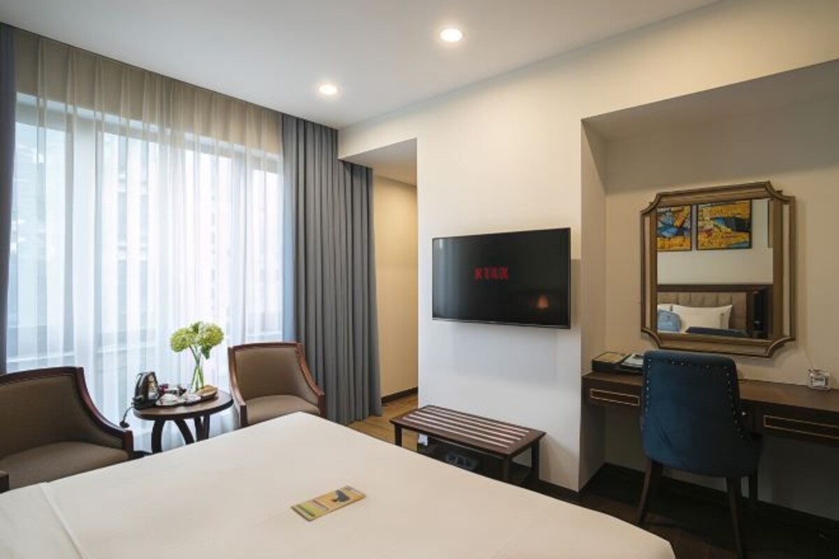 Habitación de hotel con una cama grande, un escritorio con silla, un televisor montado en la pared y una ventana con cortinas.