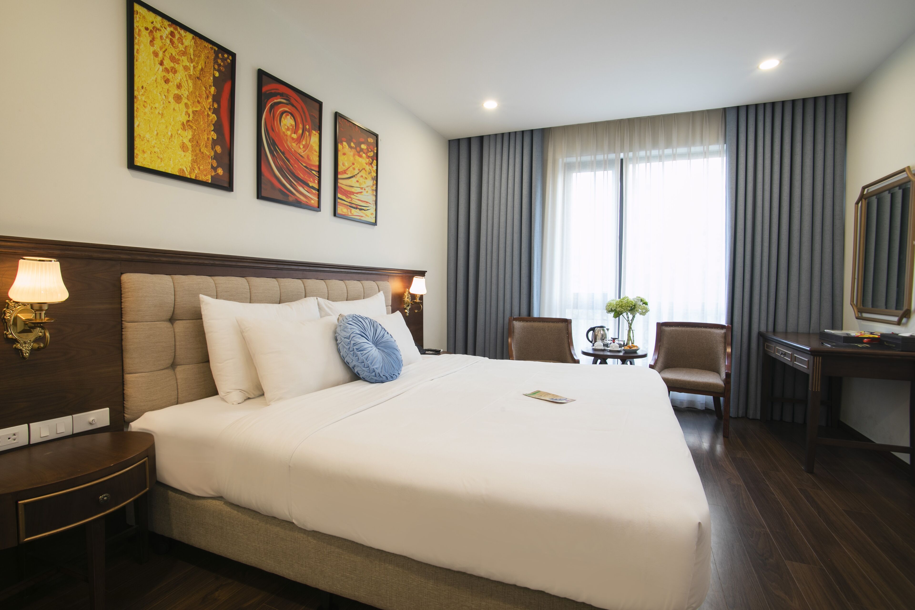A hotel room with a large bed, a desk, a chair, and two paintings on the wall.