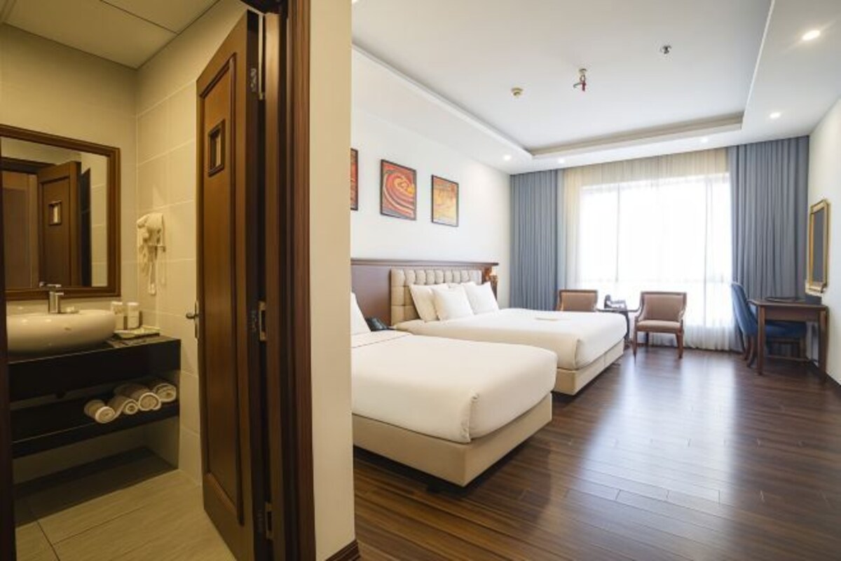Habitación de hotel con una cama grande, una cama más pequeña, un escritorio y una silla. Incluye un baño con lavamanos y espejo.