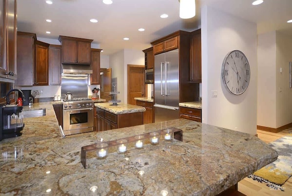 A modern kitchen with granite countertops, stainless steel appliances, wooden cabinets, and a wall clock.