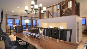 A dining area with a long wooden table, multiple chairs, a chandelier, and a staircase leading to an upper level.