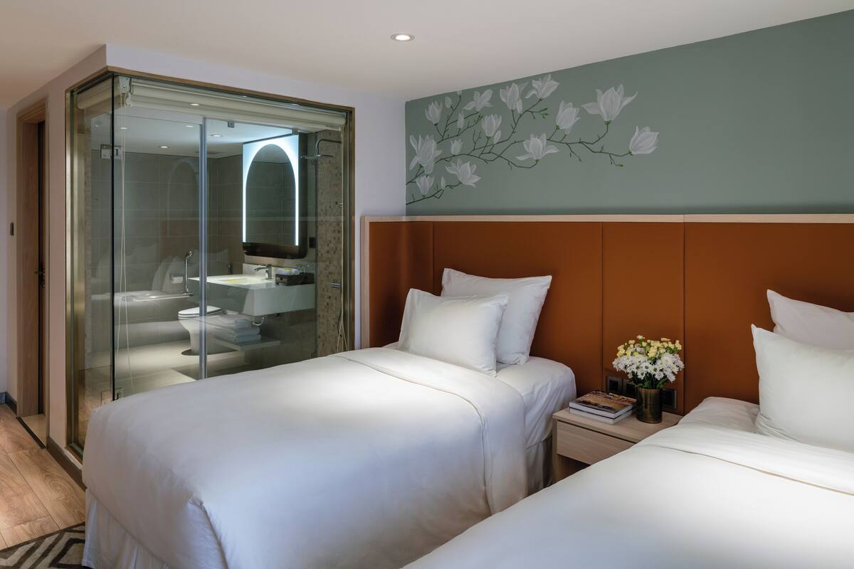 Une chambre d’hôtel avec deux lits, une douche séparée par des parois en verre, une salle de bain avec un lavabo et des toilettes, et un mur orné d’une fresque florale.