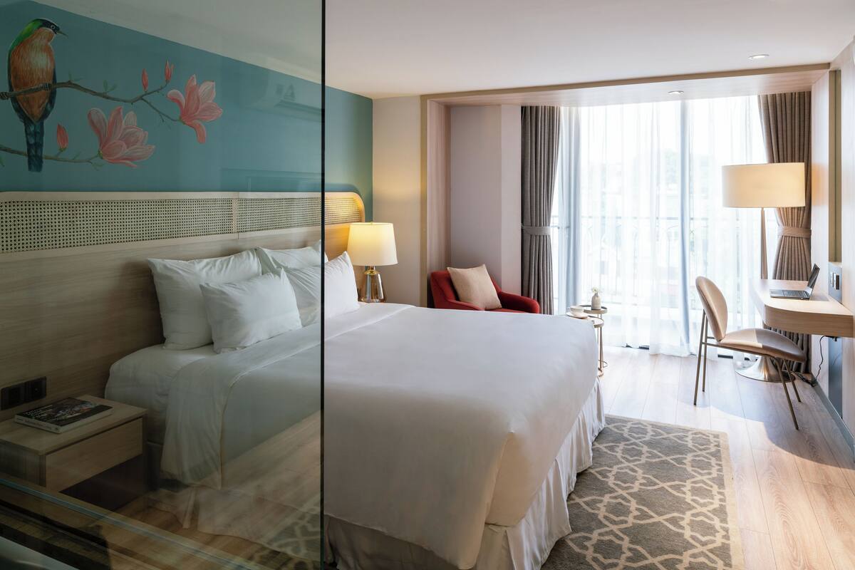 Une chambre d’hôtel avec un grand lit, un bureau avec une chaise, une petite table avec une lampe et un tableau au mur.