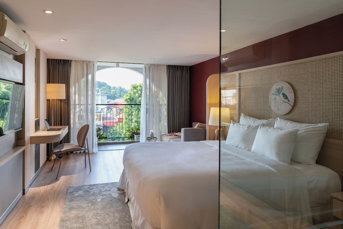 Une chambre d’hôtel moderne dotée d’un grand lit, d’un bureau avec un ordinateur portable, d’une chaise et d’un balcon offrant une vue sur la verdure.