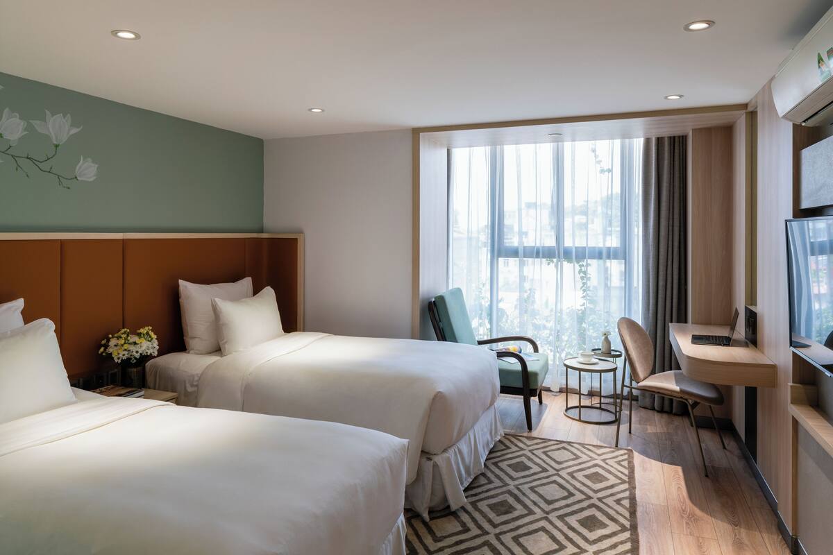 Une chambre d’hôtel avec deux lits, un bureau, une chaise, une petite table et une grande fenêtre avec des rideaux.