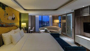 Ein modernes Hotelzimmer mit einem großen Bett, einer gebogenen Sitzecke, einem Fernseher und Blick auf die Stadt im Abendlicht.