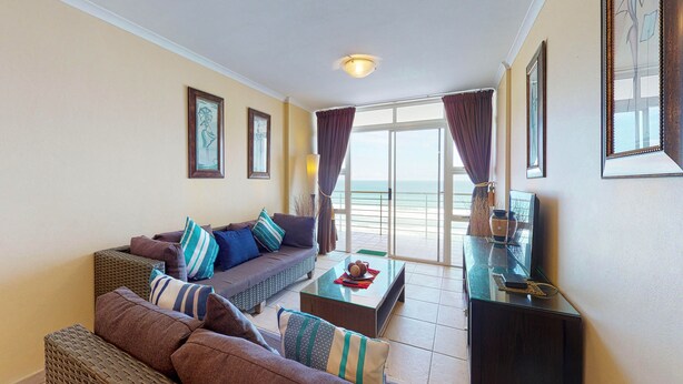 Ein Wohnzimmer mit Sofa, Couchtisch und Fernseher. Es gibt einen Balkon mit Vorhängen und Meerblick.