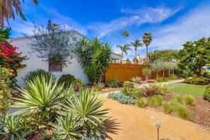 Il s’agit d’une propriété résidentielle comprenant une maison blanche, une clôture en bois et une variété de plantes, notamment des agaves et des palmiers.