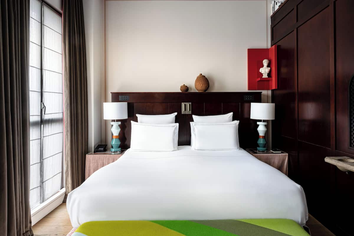 Una habitación de hotel con una cama grande, dos mesitas de noche con lámparas, un cabecero de madera y una ventana con cortinas.
