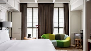 A modern hotel room with a large bed, a green and yellow armchair, a desk with a lamp, and large windows with curtains. - Experimental Marais (Paris)