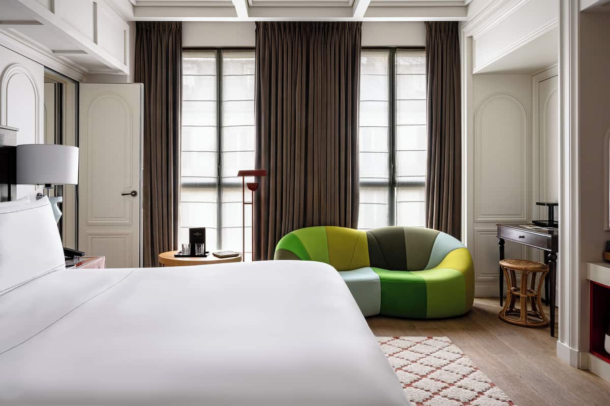 Una habitación de hotel moderna con una cama grande, un sillón verde y amarillo, un escritorio con lámpara y ventanas amplias con cortinas.