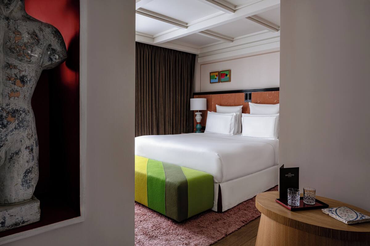 Habitación de hotel con una cama grande, una mesita de noche de madera, un reposapiés rojo y verde, una mesita de noche con lámpara y un cuadro en la pared.