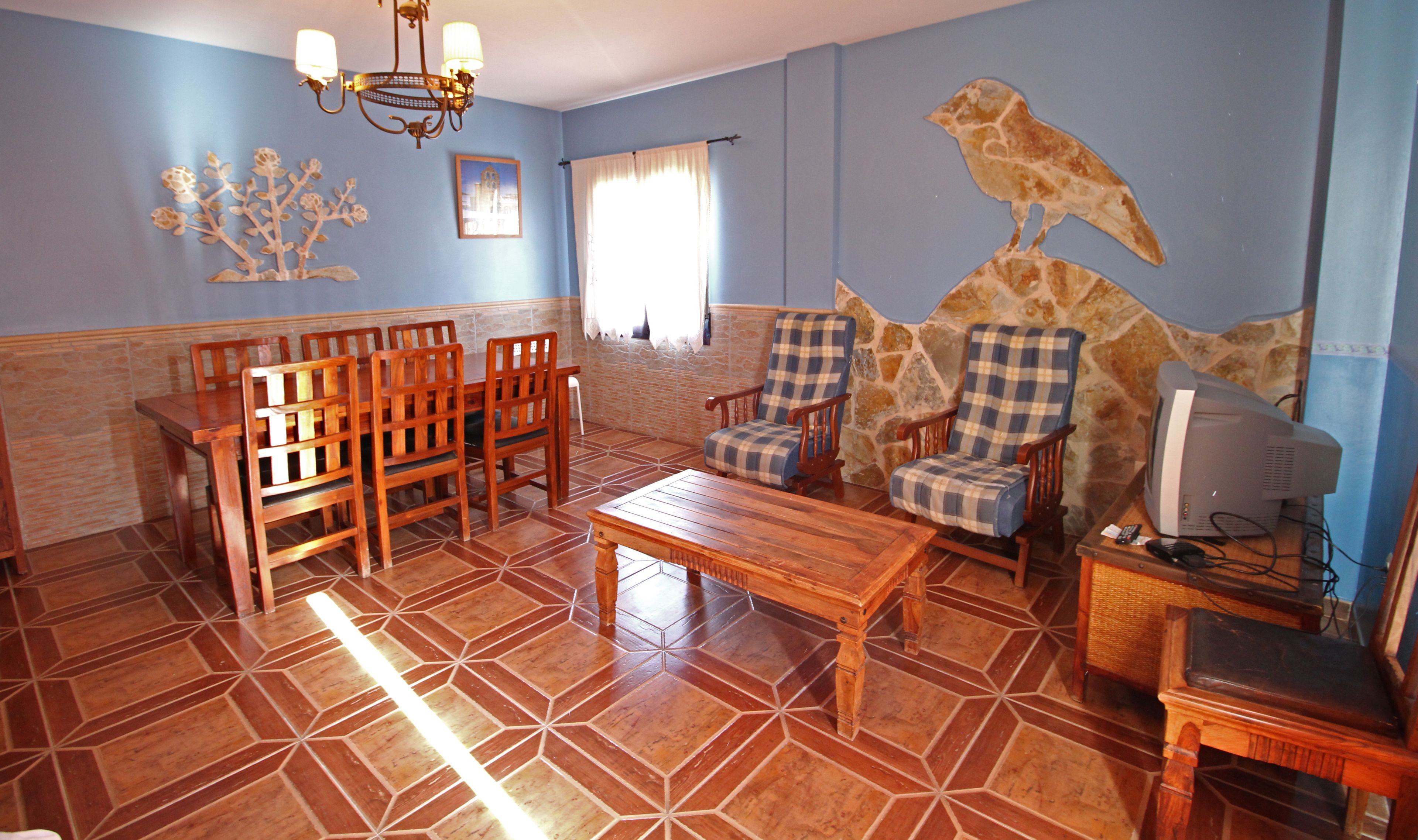 Una sala de estar con un comedor de madera, dos sillones, una mesa de centro, un televisor y una pared de piedra con un mural de aves.