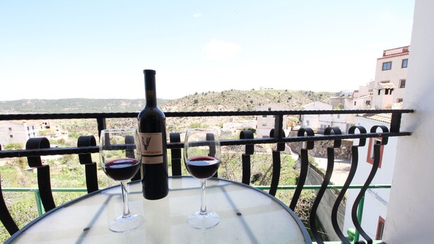 A balcony with a table holding a bottle of wine and two glasses, overlooking a hillside with buildings. - El Rincón de Piedra Apartamentos Rurales (Enguidanos)